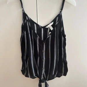 Svart randig linnetopp med knyt - Svart och vitrandig linnetopp från H&M med smala axelband, knappar framtill och snygg knytning nedtill. Luftigt material och v-ringad halsringning gör den perfekt för varma dagar. Enkel att matcha med jeans eller shorts
