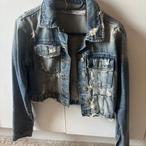 Cropped jeansjacka från Zara - Snygg croppad jeansjacka från Zara i blå denim med slitna detaljer och råa kanter. Klassisk krage, knappar framtill och bröstfickor. Perfekt för dig som gillar en edgy och trendig stil.