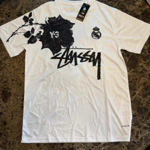 Stussy t-shirt - Vit t-shirt från Stussy Y-3 och Real Madrid med stor svart ros och Stüssy-tryck framtill. Adidas Aeroready-material som andas, kortärmad och med coola grafiska detaljer både fram och bak. Perfekt för dig som gillar streetwear och unika samarbeten.