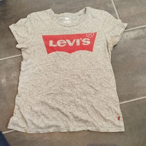Grå Levi's t-shirt med röd logga - Snygg gråmelerad t-shirt från Levi's med klassisk röd logga framtill. T-shirten har rund halsringning och korta ärmar. Perfekt för en avslappnad och cool stil. Tillverkad i mjuk bomull för skön känsla hela dagen.