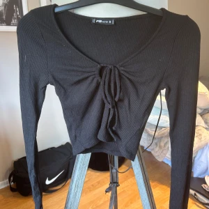 Svart ribbad croppad topp FB Sister - Svart långärmad croppad topp från FB Sister med ribbad struktur och snygg knytning framtill. Toppen har en djup rundad ringning och sitter tight. Perfekt för dig som gillar en enkel men trendig look. 🩷