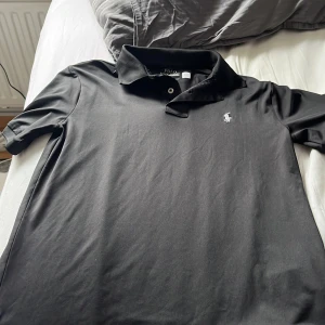 Svart pikétröja från Ralph Lauren - Klassisk svart pikétröja från Polo Ralph Lauren med vit broderad logga på bröstet. Tröjan har korta ärmar, krage och knäppning framtill. Tillverkad i ett mjukt och lätt polyestermaterial som känns skönt mot huden. Perfekt för en clean och stilren look.