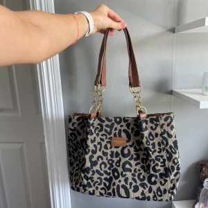 Leopardmönstrad axelväska från Patricia Nash - Snygg axelväska med leopardmönster från Patricia Nash. Väskan har bruna handtag i skinn med guldfärgade kedjedetaljer och en beige och svart canvasliknande kropp. Perfekt för dig som vill sticka ut med en trendig och djurinspirerad look.