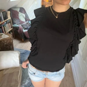 Svart volangtopp - Säljer en svart topp med volangdetaljer längs sidorna. Använd två tre gånger endast. Superfin att styla med ett par jeans.