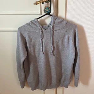 Grå hoodie med huva och dragsko - Säljer en enkel och stilren grå hoodie med huva och dragsko. Tröjan har långa ärmar och ribbade muddar vid ärmslut och nederkant. Perfekt för en avslappnad look.
