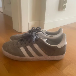 Adidas Gazelle grå/vita sneakers - Säljer ett par klassiska Adidas Gazelle sneakers i grå mocka med vita detaljer och vita ränder på sidorna. Skorna har snörning och platt sula, samt Adidas-logga på plösen och hälen. Perfekta för en stilren och sportig look.