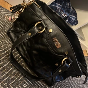 Svart quiltad axelväska från FEEL - Snygg svart axelväska från FEEL med quiltad design och guldfärgade metalldetaljer. Väskan har dubbla handtag och dragkedja upptill. Perfekt för att bära dina viktigaste saker och ger en stilren look.