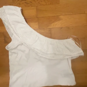 Vit oneshoulder topp med volang från Gina Tricot - Säljer en vit oneshoulder topp från Gina Tricot med bred volangkant i broderad bomull. Toppen är ärmlös och har en romantisk känsla med fina detaljer. Perfekt för varma dagar eller sommarens tillfällen.