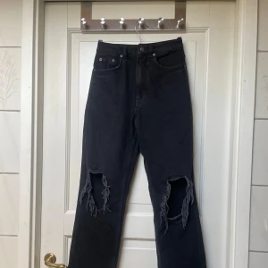 Svarta jeans med hål på knäna - Säljer ett par svarta jeans med raka ben och stora slitna hål på båda knäna. Klassisk femficksmodell med hög midja och knappgylf. Perfekta för en avslappnad och trendig look.