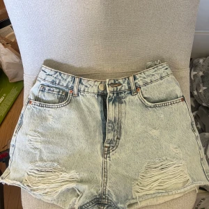 Ljusblå jeansshorts från Zara - Säljer ett par ljusblå denimshorts från Zara med hög midja och slitna detaljer framtill. Klassisk femficksmodell med knapp och dragkedja. Perfekta för sommaren och ger en avslappnad vibe.