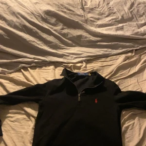 Svart tröja med dragkedja från Polo Ralph Lauren - Svart långärmad tröja från Polo Ralph Lauren med halv dragkedja och klassisk röd logga på bröstet. Perfekt för en stilren och avslappnad look. Mjuk och bekväm, passar till många tillfällen.
