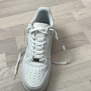 Nike Air Force 1 helvita sneakers - Klassiska helvita Nike Air Force 1 sneakers med snörning och rund tå. Skorna har en platt sula och är tillverkade i syntetmaterial med diskret Nike-logga på hälen. Perfekta för en clean och stilren look. 