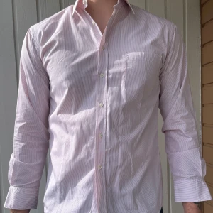 Ljusrosa randig skjorta från Stenströms - Säljer en klassisk ljusrosa skjorta från Stenströms med tunna röda ränder. Skjortan har lång ärm, bröstficka och knäppning framtill. Tillverkad i tvåfaldig super bomull för en mjuk och lyxig känsla.