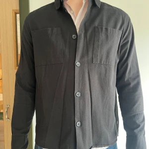 Svart overshirt från !Solid - Säljer en svart overshirt från !Solid i bomull. Jackan har två bröstfickor, knäppning framtill med stora knappar och klassisk krage. Perfekt att slänga över en t-shirt eller skjorta för en clean look.
