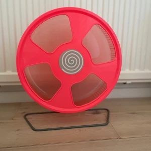 Röd hamsterhjul i plast - Säljer ett stort rött hamsterhjul i plast med transparenta fönster och spiraldekor i mitten. Hjulet står på ett stabilt metallstativ och är designat för att ge smådjur motion inomhus. Passar perfekt för hamster eller andra små gnagare.