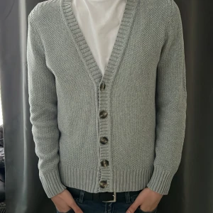 Ljusgrå stickad cardigan - Nu säljer vi en ljusgrå stickad kofta med brun/beaga knappar framtill. Koftan har lång ärm, djup v-ringning och bred ribbad kant vid ärmslut och nederkant. Perfekt för lager-på-lager och enkel att matcha med olika outfits.