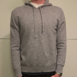 Grå hoodie i kashmir - Mjuk och stilren grå hoodie i kashmir med dragsko i huvan. Tröjan har lång ärm och ribbade muddar vid ärmslut och nederkant. Perfekt för en avslappnad och snygg look.