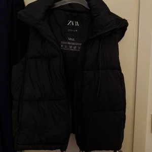 Svart dunväst från Zara - Säljer en svart dunväst från Zara med hög krage och dragkedja framtill. Västen har en puffig design och är perfekt för lager-på-lager-stil. Passar dig som vill ha en enkel och stilren look.