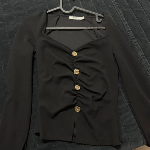 Svart topp med guldknappar från NA-KD - Snygg svart topp från NA-KD med långa ärmar och dekorativa guldknappar framtill. Toppen har en rynkad detalj längs knappraden och en v-ringad halsringning. Perfekt för att lyfta vilken outfit som helst.