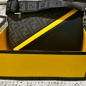 Fendi axelväska   - Snygg axelväska från Fendi i svart och grått med tydligt Fendi-mönster och en skarp gul diagonal detalj. Väskan har bred axelrem med logga och silverfärgade metalldetaljer, samt dragkedja. Perfekt för dig som vill sticka ut med stil.