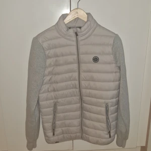 Ljusgrå cardigan från Koroshi - Säljer en ljusgrå pufferjacka från Koroshi med quiltad front och rygg samt mjuka ärmar. Jackan har dragkedja framtill och två fickor med dragkedja. Perfekt för kyliga dagar och enkel att matcha med olika outfits.