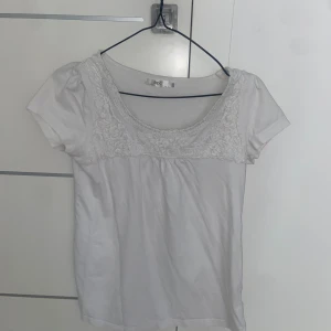 Vit blus med spetsdetaljer - Säljer en söt vit blus med korta ärmar och vacker spets längs den breda, rundade ringningen. Perfekt för en fräsch och stilren look. Blusen är lätt och har en feminin känsla.