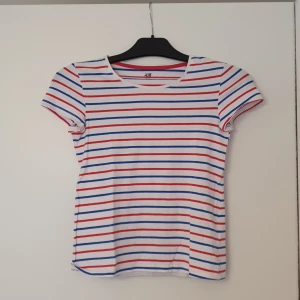 Randig t-shirt från H&M - Säljer en vit t-shirt från H&M med röda och blå horisontella ränder. T-shirten är i ekologisk bomull och har en klassisk passform med rund hals och korta ärmar. Tröjan har storleken 146/152