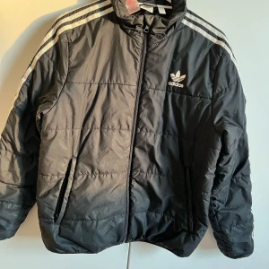 Svart pufferjacka från Adidas - Svart pufferjacka från Adidas med klassiska vita ränder på axlarna och logga på bröstet. Jackan har dragkedja framtill, hög krage och två fickor med dragkedja. Tillverkad i syntetmaterial.