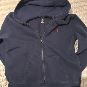 Marinblå hoodie från Polo Ralph Lauren - Säljer en marinblå hoodie från Polo Ralph Lauren med dragkedja och huva. Tröjan har en röd broderad logga på bröstet och två fickor framtill. Perfekt för en avslappnad stil. mycket populär! Bra pris!?!?! och nästan helt ny 