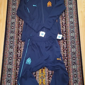 Marinblå träningsjacka från Puma x Olympique de Marseille - Säljer en marinblå träningsjacka och byxa från Puma i samarbete med Olympique de Marseille. Jackan har huva, dragkedja framtill, broderad klubbmärke och Puma-logga samt detaljer i orange och ljusblått. Perfekt för fotbollsentusiasten! Byxan är i storlek XL och jackan är i L
