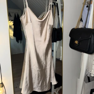 Champagnefärgad satin klänning  - ALDRIG ANVÄND!, köpt i höstas.  Elegant champagnefärgad mini klänning med smala och justerbara axelband. Klänningen har en slät yta och är perfekt för en stilren look. Den har en figurnära passform som framhäver silhuetten. Prislapp kvar.