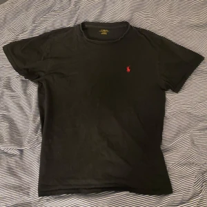 Svart t-shirt från Polo Ralph Lauren - Klassisk svart t-shirt från Polo Ralph Lauren med röd broderad logga på bröstet. T-shirten har rund hals och korta ärmar. Perfekt basplagg med stilren design och skön passform.