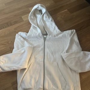 Vit hoodie med dragkedja - Säljer en klassisk vit hoodie med dragkedja och huva. Tröjan har långa ärmar, två fickor framtill och är tillverkad i ett mjukt material som passar perfekt för en avslappnad stil.
