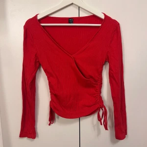 Röd långärmad omlottopp med knyt - Röd omlottopp med v-ringning och långa ärmar. Toppen har ribbad struktur och knyt i sidan som ger en snygg detalj. 