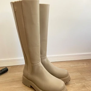 Beiga knähöga stövlar  - Säljer ett par snygga beige höga boots med chunky sula. Skorna har en stilren design utan synliga detaljer och är perfekta för dig som vill ha ett par trendiga boots till vardags. De är aldrig använda och de har storlek 40. 