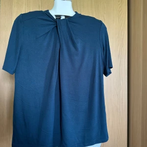 Mörkblå topp med knutdetalj från Stockh LM - Säljer en mörkblå topp från Stockh LM med korta ärmar och snygg knutdetalj vid halsen. Modellen är rak och enkel, perfekt för en stilren look. Materialet känns mjukt och bekvämt.