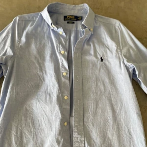 Ljusblå skjorta från Polo Ralph Lauren ( stl 16) junior - Klassisk ljusblå skjorta från Polo Ralph Lauren med broderad logga på bröstet. Skjortan har lång ärm, knappar framtill och button-down krage. Perfekt för en stilren och avslappnad look.