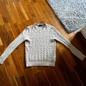 Biege kabelstickad tröja från Morris - Säljer en klassisk biege kabelstickad tröja från Morris. Tröjan har rund halsringning och långa ärmar med ribbade muddar och nederkant. Perfekt för en stilren och tidlös look. Loggan har lossnat på ett hörn med inget man tänker på