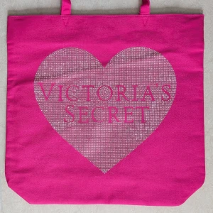 Rosa tygväska från Victoria's Secret - Säljer en snygg rosa tygväska från Victoria's Secret med stort hjärtmotiv och logga på framsidan. Väskan har två handtag och är rymlig med svart innerfoder. Perfekt för att bära med sig allt man behöver. Tygväskan har aldrig använts 