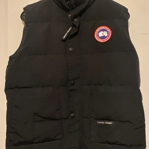 Svart dunväst från Canada Goose - Säljer en svart Canada Goose dunjacka. Den här västen är perfekt för att hålla dig varm under kyliga dagar utan att kompromissa med stilen. Den har den ikoniska Canada Goose-logotypen och är i utmärkt skick. Perfekt för lager-på-lager eller som ett fristående plagg. Missa inte chansen att äga denna lyxiga och högkvalitativa väst! Den är storlek s 