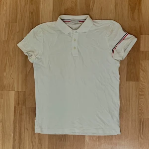 Vit piké från Moncler med röd och svart detalj - Säljer en klassisk vit pikétröja från Moncler med knappar framtill och broderad Moncler-logga på ärmen. Snygga röd och svart ränder på krage och ärm ger en sportig touch. Perfekt för dig som gillar stilrena plagg.