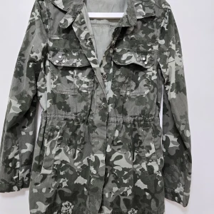 Grön camouflagejacka med knappar - Säljer en grön camouflagejacka med långa ärmar och knappar framtill. Jackan har två bröstfickor med dekorativa nitar och dragsko i midjan för justerbar passform. 