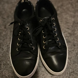 Svarta sneakers från Vagabond - Svarta sneakers från Vagabond med vita sulor och svarta skosnören. Skorna har guldfärgade öljetter och detaljer i blankt material vid hälen. Klassisk rund tå och platt sula, tillverkade i skinn.