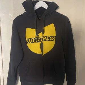 Wu-Tang svart hoodie med gul logga - Svart hoodie med stor gul Wu-Tang Clan-logga på bröstet. Klassisk huva med dragsko och känguruficka framtill. Mjuk insida och normal passform, perfekt för dig som gillar streetwear och hiphopstil.