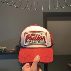 Röd och vit keps med broderad patch - Säljer en röd och vit keps med broderad patch framtill där det står 'The Indian Skeleton Dime'. Kepsen har mesh på sidorna och bak, samt justerbar knäppning. Perfekt för dig som gillar streetwear eller vill ha en cool accessoar.