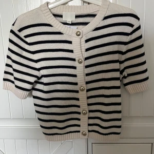 Randig kortärmad kofta med pärlknappar - Säljer en söt stickad med ränder. Koftan har korta ärmar, rund halsringning och dekorativa pärlknappar framtill. Ny med prislapp