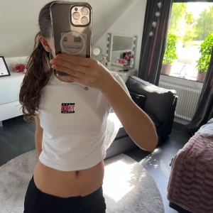 Vit croppad t-shirt från Tommy Hilfiger - Säljer en vit croppad t-shirt från Tommy Hilfiger med logga på bröstet. T-shirten har korta ärmar och rund halsringning. Perfekt för en avslappnad och stilren look.