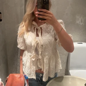 Vit blus med knyt och volang - Säljer en söt vit blus med knyt framtill och volangdetaljer. Blusen har korta puffärmar och strukturmönster i tyget. Perfekt att styla med jeans eller kjol för en trendig look.