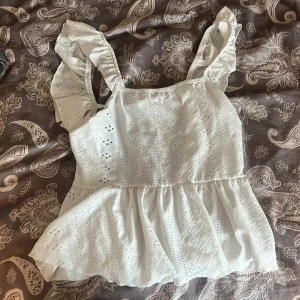 Vit volangtopp med broderade detaljer - Söt vit topp med volangaxelband och broderade hålmönster.  Toppen har en peplum-liknande nederdel och knytning i ryggen för extra detalj. Perfekt för sommardagar och ger en romantisk känsla.