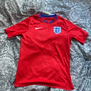 England röd fotbollströja från Nike - Säljer en röd England fotbollströja från Nike med blå detaljer i kragen och det klassiska landslagsmärket på bröstet. Tröjan är kortärmad och tillverkad i Dri-FIT material som andas. Perfekt för fotbollsträning eller att bära till matchen!